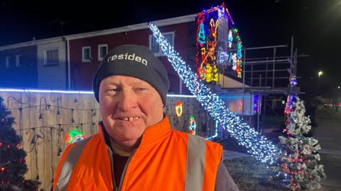 Head and shoulders image of Stan Yanetta standing in front of his illuminated house. Mr Yanetta is looking into the camera and wearing a black woolly hat and orange hi-vis jacket. The gable end of his house is lit up, as is a wooden garden fence. A Christmas tree with fairly lights can be seen over his shoulder.