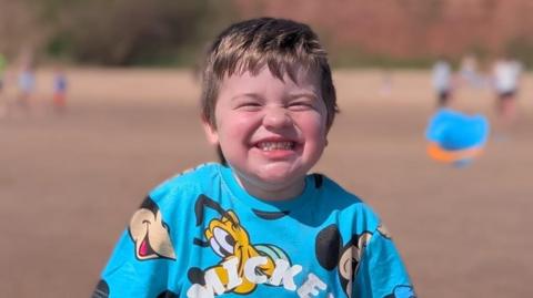 Louie smiles on a beach. He wears a bright blue Mickey Mouse T-shirt, and has short blond hair.