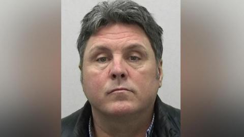 Mugshot of Nellis. He has a chubby face and is clean shaven with short think black and grey hair.