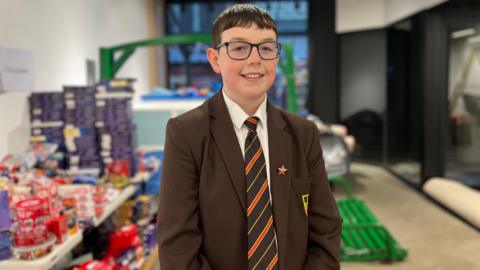 Joseph stands in front of a table full of selection boxes and boxes of sweets. He has short, dark hair and wears dark-rimmed glasses. He wears a brown school blazer and a brown and orange striped tie