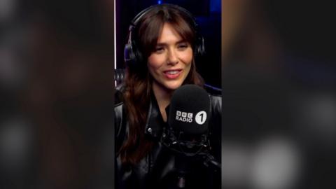 A woman, Elizabeth Olsen, with long dark hair behind a Radio 1 microphone.
