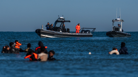 French police vessels look on as migrants wade into the sea to try and board a dinghy to cross the English Channel