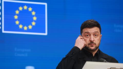 Volodomyr Zelensky, a man with short dark hair and beard who is wearing a black shirt, stands at a lectern in front of a blue background with an EU flag on it.