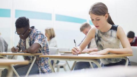 Students, sitting an exam in a hall. Image shows five students sitting at exam-style desks, holding pens and reading or writing.