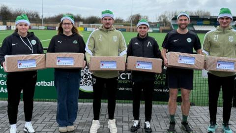 Two women and four men dressed in sports kit are standing on front of a football pitch. They are all wearing green santa hats and holding boxes wrapped in Christmas paper, with greetings tags on them.
