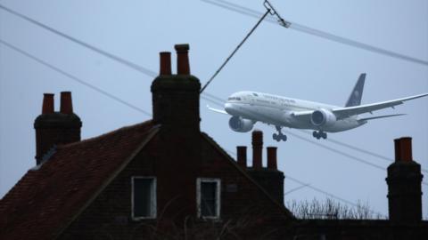 A plane approaching Heathrow Airport, flying low over residential rooftops with chimneys and power lines visible.