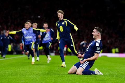 Scotland's Kenny MacLean is on his knees on the Hampden pitch as he celebrates scoring his goal against Denmark. Scotland substitutes run towards him to congratulate him.
