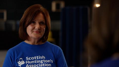 Gillian McNab - woman with mid-length red hair, wearing a blue t-shirt which reads "Scottish Huntington's Association"