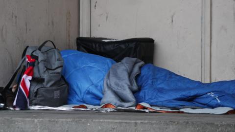 A person in a blue sleeping bag. They have a grey rucksack next to them with a flag attached to it.