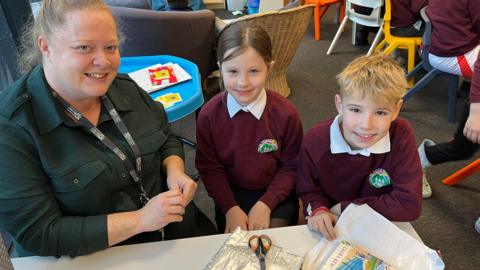 Two children with maroon tops and white collars sit beside a woman in green top with tied back hair. In front of them is a desk with half wrapped books