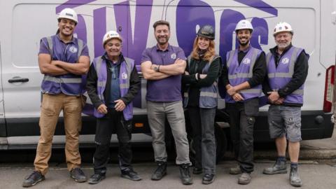 Llun o dîm DIY SOS yn sefyll yn edrych at y camera ac yn gwenu gyda fan gwyn tu ôl iddyn nhw. 6 person sydd, 5 dyn a 3 menyw. I'r chwith ma dyn tal gyda het galed wyn ar ei ben. Mae'n gwisgo crys-t porffor a throwsus melyn. Nesa iddo fe mae dyn arall sy'n gwisgo het galed wyn a siaced du. Nesa mae dyn gyda gwallt brown, crys-t porffor a throwsus llwyd. Yna mae menyw gyda het galed du, siaced du a throwsus du. Nesa iddi hi mae dau ddyn arall. Mae gan y ddwy het galed wyn a siwmper du.