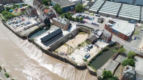 An aerial view of a dry dock. There are a number of buildings and a large concrete base. There is a body of water in front of the dock and the muddy river bank is visible.