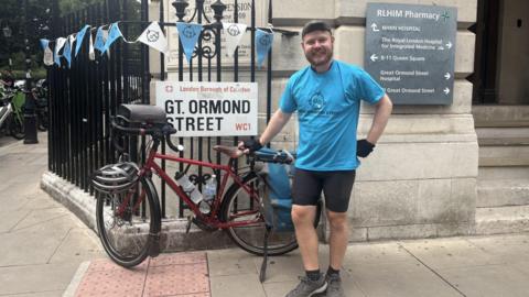 A man looks at the camera, with his right hand on his bike and left hand on his hip. He is wearing a Great Ormond Street Hospital Charity t-shirt and is surrounded by charity bunting, stood outside of the hospital in London.
