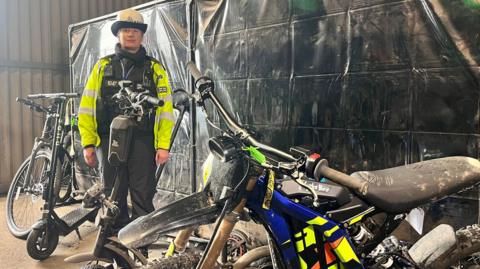 A female police officer dressed in a black uniform, yellow hi-vis vest and white hat stands in a warehouse surrounded by electric bikes and scooters. Behind her is a black screen. The bike closest to the camera resembles a scrambler motorcycle, with a large saddle and colourful cowling.