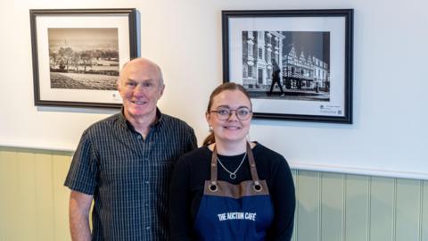 Joe Anderson, a man wearing a dark-coloured shirt, and cafe manager Emma Dermody, a woman wearing a black top and a dark blue apron, are standing in front of two monochrome images on a wall which is white with green wood panelling on the lower half.