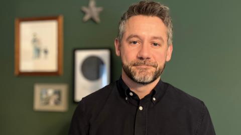 Kieren Jennings looking into the camera with a straight face. He has short, grey and brown hair and beard. He is wearing a black shirt. He is standing in front of a green wall with various frames in the left corner.
