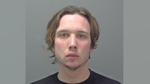 A mugshot head and shoulders image of Elliott Mallett. He is wearing a black t-shirt and standing against a grey wall. He is staring into the camera.