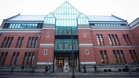 A general view of Minshull Street Crown Court in Manchester. The court is a two-storey red brick building with a glass-panel frontage. People can be seen walking past the entrance on a grey day.