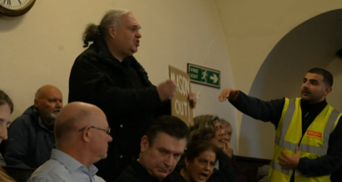 A man holds a cardboard sign saying "Mason out!" while standing up. Other people around him sit down and a man in a hi-vis vest gestures towards him.