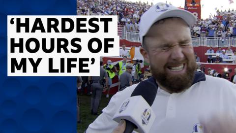 Shane Lowry, wearing a white polo shirt and white cap, grimaces whilst being interviewed