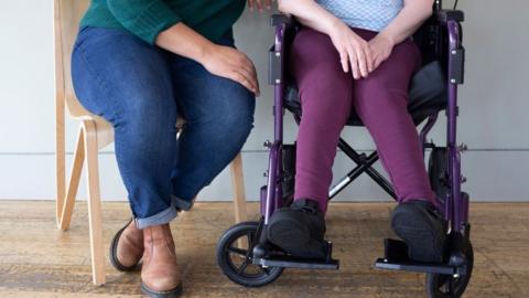 Two people sitting close together, One of the women has quadriplegic cerebral palsy and is a wheelchair user. The woman sat on the left is wearing a green jumper, blue jeans and brown boots, sat on a wooden chair, the person using the wheelchair has purple trousers and a blue top. They are pictured from the waist down.