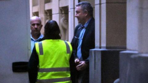 A middle-aged man in handcuffs is led out of a building by a woman in a fluorescent jacket