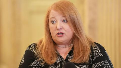 Naomi Long has long red hair and is wearing a black and white top.