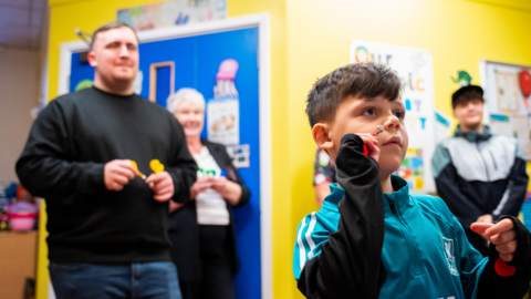 In a hospital ward, a dark-haired boy wearing a blue/green tracksuit top is poised to throw a magnetic dart. Behind him is Luke Littler, waiting his turn with more darts. Another adult stands behind smiling in front of a bright blue door in the yellow-walled room. An older boy watches from the other side of the child, and smiles.