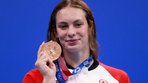 Penny Oleksiak stands against a blue background and smiles as she holds up a bronze medal at the 2020 Tokyo Olympic Games. She is wearing a red and white jacket.