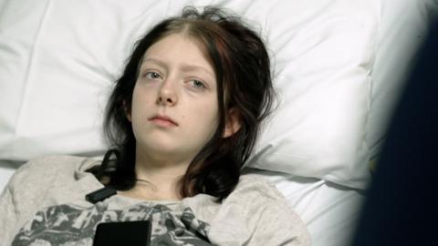Paige who is 19 is lying on a hospital bed resting her head on a pillow. She looks ill. She has dark hair and is wearing a T-shirt.