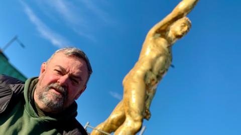 A man in a green hoodie reading 'Bulldog Forge' posing in front of a large gold statue of a roman male gladiator. The gladiator has has curly short hair on it's head and a fist raised in the air. It is naked and male but is blurred into the background in this shot. Blue sky and rooftops can be seen behind the big statue.