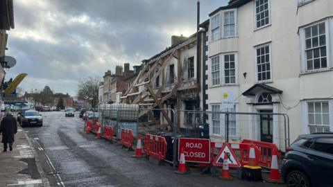 A fire damaged building being propped up by wooden beams. A white building next door is also cordoned off. The cordon takes up much of the road leaning to just one lane being usable