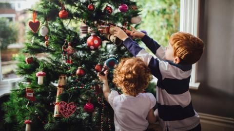 A young red head boy and his sister are decorating the Christmas tree in their home. He is reaching up and putting a bauble onto the tree while his sister tries to help.