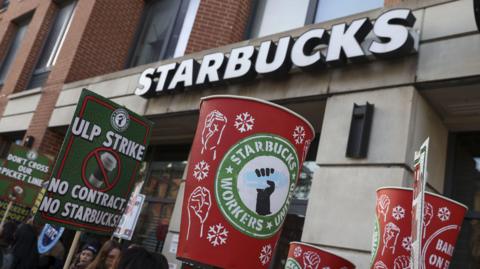 Demonstrators hold signs reading "ULP Strike" and "No Contract, No Starbucks!" in front of a Starbucks store.