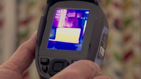 A man's hand holds a small thermal imaging camera which is made out of grey plastic and has a number of buttons on the bottom of it. The image in the middle of the camera is picking up on the area of the room it is pointed at and shows the warm areas in yellow and the cold areas on blue.