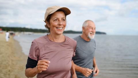 A middle-aged woman and man are running along a beach, with sea and a cloudy sky in the background