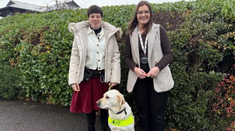 Blind women with a guide dog in front of her. There's green shrubbery behind. She's stood next to a women in glasses and they are both smiling at the camera.