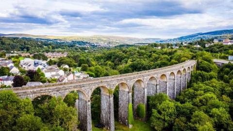 Cefn Viaduct with Merthyr Tydfil in the background - 13 large brick arches belonging to the viaduct can be seen in the foreground, surrounded by trees, houses and rolling hills.