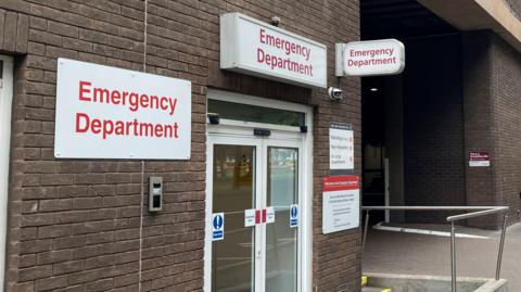 A picture of the front doors of the Emergency Department in St Helier, Jersey. The hospital building is made up of brown bricks, and the doors are glass.