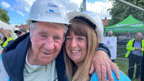 A man and woman both wearing white hard hats smiling. The man has his arm around the woman and their heads are touching. They're outdoors near a fence, with other people in safety gear and a green tent in the background.