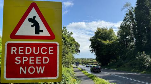 A large road safety sign on the side of the A5 in County Tyrone which reads: "Reduce speed now". The yellow, red and white sign warns motorists of a left-hand bend ahead with three off-slips, one before, one after and one in the middle of the bend. A number of vehicles are driving along the road on a sunny day.
