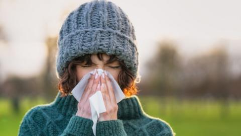 A woman outside in a grey wool hat and green jumper blows her nose on a tissue.