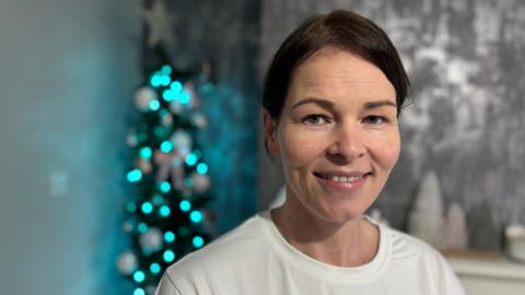 A woman with brown hair and a white t shirt smiles in front of a christmas tree