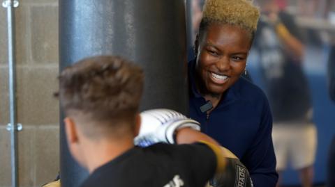 A child with short brown hair seen from behind with a white boxing glove on that is hitting a black punchbag. Nicola is leaning close to the punchbag with a lovely grin, looking at the child.