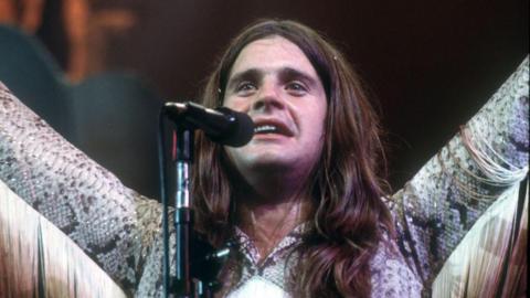 A long-haired singer in front of a microphone with snake-skin outfit with tassels on the arms