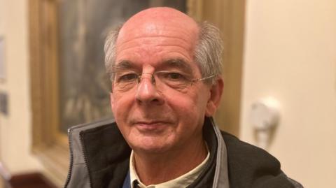 A man with balding grey hair and wearing glasses looks toward the camera. He is wearing a grey coat, white shirt and blue jumper.