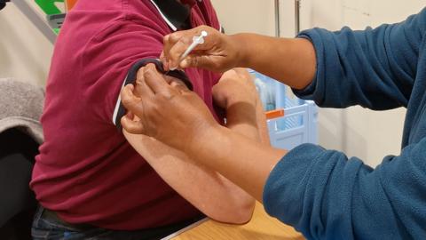 A man is sat on the left with his arm on the table. He is wearing a red polo shirt and someone is putting a vaccine in his upper arm. A person is on the right who is giving the vaccine, and is wearing a blue jumper.