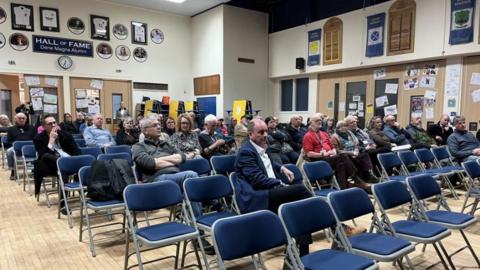 The picture has been taken in a school hall. Blue backed chairs are in neat rows. A number of chairs at the front are empty but there are people sitting further back listening to what is been said.