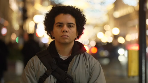 Keira Bell stands in front of a dark, black curtain, wearing a black hoodie with red and yellow designs near the elbows. She has dark, curly, short hair, dark brown eyes and wears earrings in her ears.
