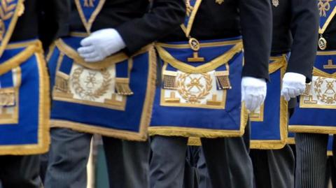 Masons march in a procession. Pictured are men from waist height wearing white gloves and wearing the Freemasons coat of arms embraided with gold thread on a royal blue background